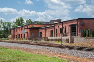 A row of brick industrial buildings with large windows, set alongside train tracks under a partly cloudy sky.