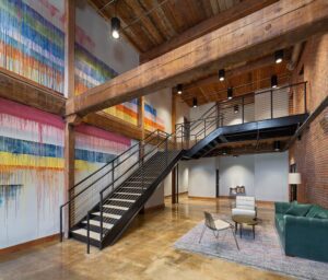 Modern loft interior with colorful wall art, metal staircase, exposed wooden beams, green sofa, chairs, and a rug on concrete floor.