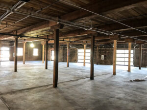 An empty industrial warehouse interior with concrete floors, wooden beams, and exposed ceiling pipes.