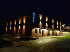 A brick building at night, with illuminated wall lights and a vertical sign reading “MARKLEY.” Sidewalks and grass area in the foreground.