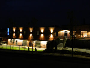 A brick building at night with illuminated exterior lights and a well-lit grassy area in front.