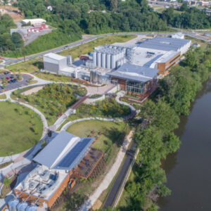 Aerial view of a riverside facility with solar panels, landscaped gardens, and parking, surrounded by trees and adjacent roads.