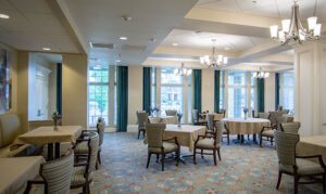 A well-lit dining area with round tables, upholstered chairs, chandeliers, and large windows with drapes.