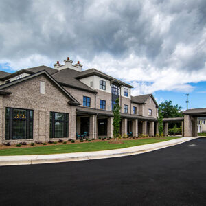 A modern, three-story brick building with large windows sits under a cloudy sky, surrounded by a paved driveway and landscape.