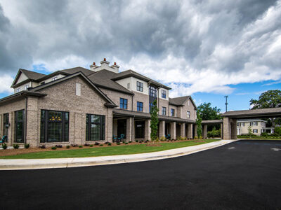 A modern, three-story brick building with large windows sits under a cloudy sky, surrounded by a paved driveway and landscape.