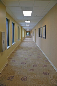 A long, empty hallway with patterned carpet, framed pictures on the walls, and windows on one side. Ceiling lights are on.