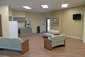 A waiting room with couches, a wall-mounted TV, and a kitchenette area featuring a refrigerator and cabinets.