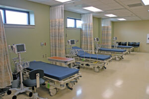 A row of empty hospital beds with monitors and curtains in a medical ward, featuring a light green and beige color scheme.