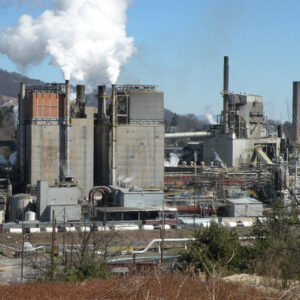 Industrial plant with smoke stacks and steam against a hilly backdrop, surrounded by trees and infrastructure.