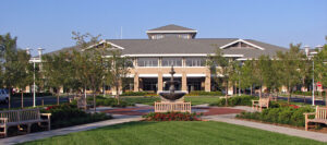 Large building with a central entrance, surrounded by trees and benches, with a fountain in front and clear blue sky above.