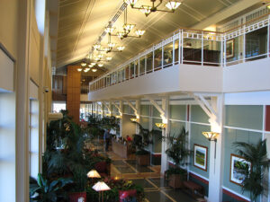 Spacious modern lobby with high ceiling, large windows, green plants, and hanging lights. Mezzanine overlooks seating area.