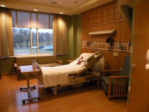 Hospital room with an empty adjustable bed, a side table, armchair, sofa, and a window with a view of trees and parked cars.