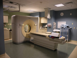 A clean, well-lit room with a large CT scanner beside an adjustable patient table. Various medical equipment is visible around.