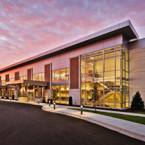 Modern building with large glass windows, surrounded by a paved driveway, against a vibrant dusk sky.