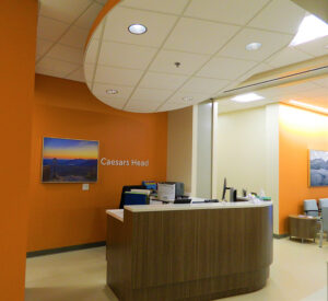 Reception desk with a computer and chairs in front of an orange wall marked "Caesars Head." Framed artwork on the wall.