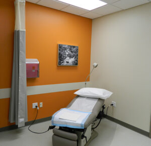 Medical examination room with an orange accent wall, an exam table, a wall-mounted light, and a landscape photo.