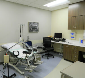 Medical examination room with a gynecological chair, computer, and cabinets under fluorescent lighting.