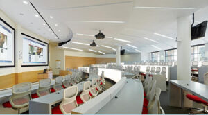 A modern lecture hall with tiered seating, white and red chairs, multiple screens, and ceiling-mounted projectors.