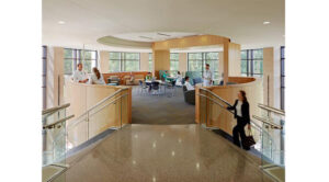 A modern hospital lobby with glass railings, people in medical uniforms, seated visitors, and large windows providing natural light.