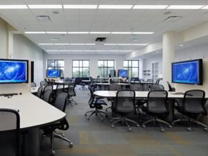 A modern classroom with multiple tables, chairs, and screens displaying blue images, set in a bright room with large windows.