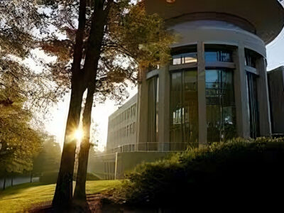 A modern building with large windows is surrounded by trees, illuminated by sunlight in the early morning or late afternoon.