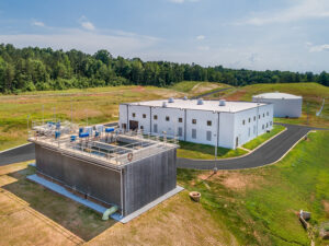 Aerial view of a white industrial building next to a large rectangular structure, surrounded by greenery and trees.