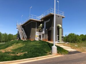 Industrial facility with metal staircase, pipes, and a flat roof, set on grassy terrain under a clear blue sky.