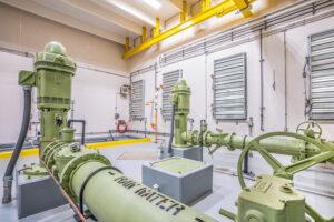 A water treatment facility room with green machinery, pipes, and pumps. White walls and a yellow overhead beam are visible.