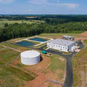 Aerial view of a wastewater treatment plant with a white cylindrical tank, rectangular building, and two settling ponds.