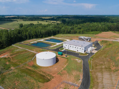 Aerial view of a wastewater treatment plant with a white cylindrical tank, rectangular building, and two settling ponds.