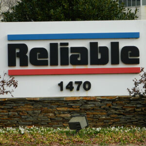 Sign with the word "Reliable" and the number "1470" on a stone base, surrounded by shrubs and parked cars in the background.