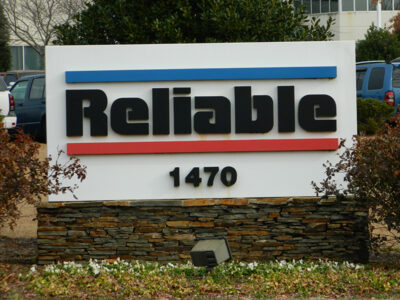 Sign with the word "Reliable" and the number "1470" on a stone base, surrounded by shrubs and parked cars in the background.