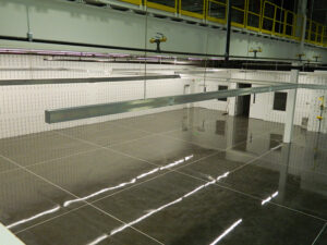 Industrial facility interior with reflective flooring, white tiled walls, overhead pipes, and ceiling beams.