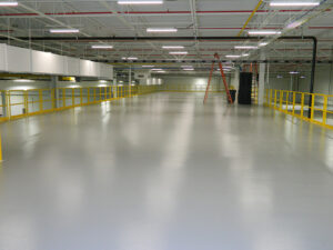 Spacious industrial warehouse interior with empty gray floor, yellow safety rails, and overhead lighting.