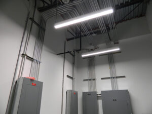 A room with gray electrical panels, metal conduits on the walls and ceiling, and bright fluorescent lighting.