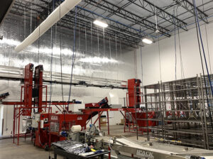 Industrial warehouse with red machinery, conveyor belts, and metal shelving under a high ceiling with exposed beams.