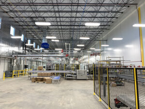 Industrial warehouse interior with machinery, equipment, and stacked boxes. High ceiling and fluorescent lighting are visible.