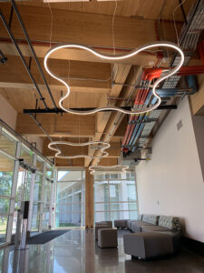 Modern lobby with abstract ceiling lights, glass walls, wooden ceiling, and gray seating area.