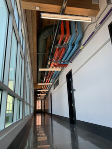 A long hallway with polished floors, large windows on one side, and colorful exposed pipes on the ceiling.