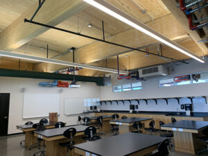 Modern classroom with wooden ceiling beams, rows of tables and chairs, large windows, and lab equipment on shelves.