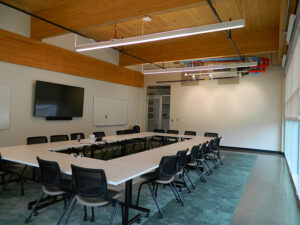 Empty conference room with U-shaped white tables, black chairs, a large wall-mounted screen, and overhead fluorescent lights.