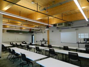 Empty classroom with rows of white desks, black chairs, a projector, whiteboards, and wooden ceiling with exposed pipes.