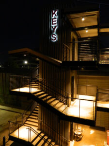 Outdoor staircase at night, illuminated with warm lights, leading to an entrance. A vertical "KEYS" sign is displayed above.