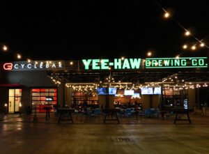 Outdoor seating area at night under string lights; signs for CycleBar and Yee-Haw Brewing Co. are visible.