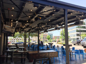 Outdoor dining area with blue chairs, wooden tables, and string lights under a metal roof. Buildings and parked cars in the background.