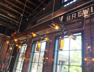 Industrial-style lighting fixture made from pipes and bottles hangs from a wooden beam in a brick-walled space.