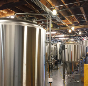 Rows of large stainless steel tanks inside a brewery with exposed wooden beams and overhead lighting.