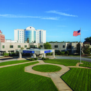 Wide view of a hospital complex with manicured lawns, an American flag, and a multi-story building in the background.