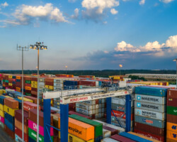 Industrial Projects Stacks of colorful shipping containers at a busy port, with cranes and lighting towers under a partly cloudy sky.