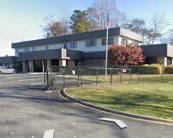 Office building with a fenced parking lot, surrounded by autumn trees. A few cars are parked, and there's a sign near the entrance.